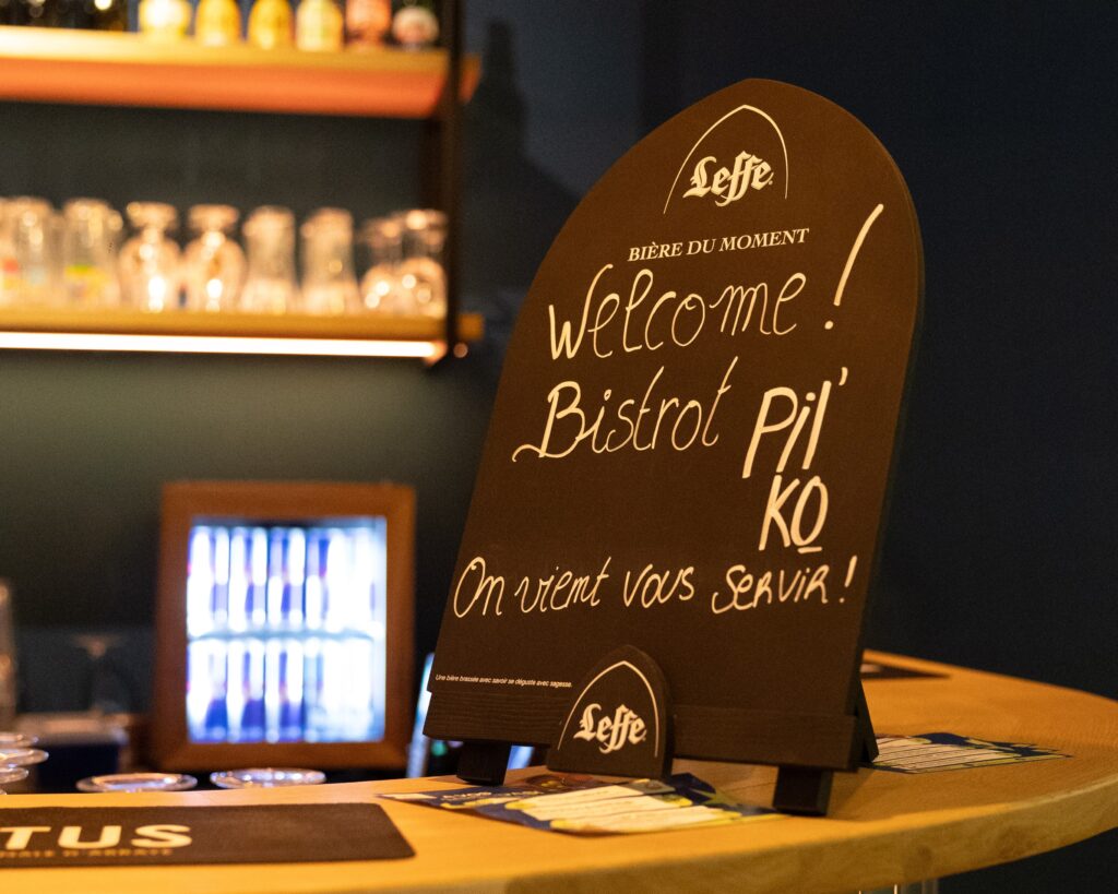 Cette image met en avant l’ambiance chaleureuse du Bistrot Pilko du groupe Tero, au Justine Henin Sports Hub. Au premier plan, un panneau noir sur un comptoir en bois affiche un message écrit à la main : "Welcome! Bistrot Pilko - On vient vous servir!", accompagné du logo Leffe, suggérant une bière du moment. En arrière-plan, des étagères illuminées présentent une sélection de verres et de bouteilles, ajoutant une touche conviviale et authentique à l’espace. Un frigo rempli de boissons est également visible, renforçant l’atmosphère accueillante de ce lieu où se mêlent sport et détente.