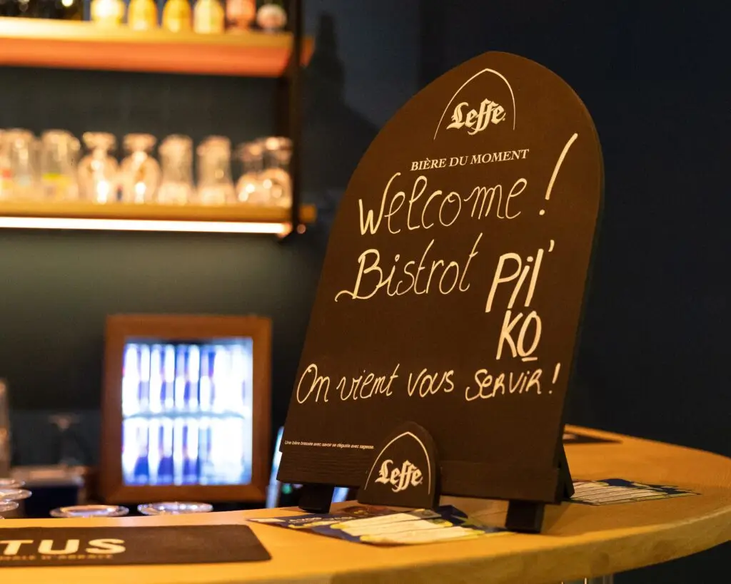 Cette image met en avant l’ambiance chaleureuse du Bistrot Pilko du groupe Tero, au Justine Henin Sports Hub. Au premier plan, un panneau noir sur un comptoir en bois affiche un message écrit à la main : "Welcome! Bistrot Pilko - On vient vous servir!", accompagné du logo Leffe, suggérant une bière du moment. En arrière-plan, des étagères illuminées présentent une sélection de verres et de bouteilles, ajoutant une touche conviviale et authentique à l’espace. Un frigo rempli de boissons est également visible, renforçant l’atmosphère accueillante de ce lieu où se mêlent sport et détente.