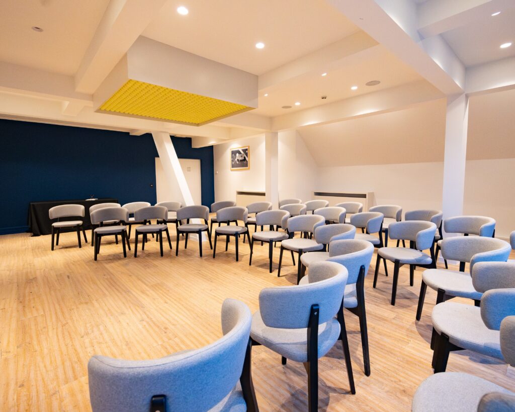 Cette image représente une des salle de séminaire du Lime, espace de réunion du groupe Tero situé au sein du Justine Henin Sports Hub, un club dédié au tennis et au padel. L’espace est lumineux et moderne, avec un sol en bois clair et un mur bleu foncé apportant une touche d’élégance. Les chaises confortables, au design contemporain avec un revêtement en tissu gris et des pieds en bois noir, sont disposées en arc de cercle, favorisant l’interaction. Un élément distinctif du lieu est la décoration suspendue au plafond, constituée de balles de tennis, un clin d’œil à l’univers du club. Cet espace polyvalent est idéal pour des réunions, séminaires et événements professionnels dans un cadre sportif et inspirant.