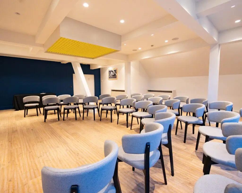 Cette image représente une des salle de séminaire du Tero Eventhouse at Lime, espace de réunion situé au sein du Justine Henin Sports Hub, un club dédié au tennis et au padel. L’espace est lumineux et moderne, avec un sol en bois clair et un mur bleu foncé apportant une touche d’élégance. Les chaises confortables, au design contemporain avec un revêtement en tissu gris et des pieds en bois noir, sont disposées en arc de cercle, favorisant l’interaction. Un élément distinctif du lieu est la décoration suspendue au plafond, constituée de balles de tennis, un clin d’œil à l’univers du club. Cet espace polyvalent est idéal pour des réunions, séminaires et événements professionnels dans un cadre sportif et inspirant.