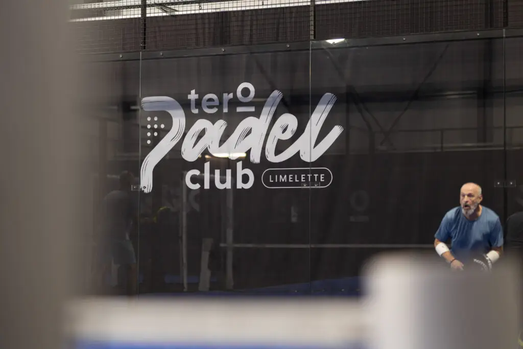 Cette image met en avant une scène de jeu sur un terrain de padel au sein du Tero Padel Club Limelette. Au premier plan, le logo du club est imprimé en blanc sur la paroi vitrée, renforçant l’identité du lieu. En arrière-plan, un joueur vêtu de bleu est en pleine action, raquette à la main, concentré sur l’échange. Au premier plan, un élément de la structure du terrain est visible mais flou car il est très proche de l'objectif, ce qui ajoute de la profondeur à l’image. L’éclairage intérieur et les structures métalliques du court créent une atmosphère immersive, reflétant l’énergie et la convivialité de ce sport en plein essor