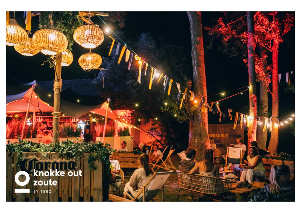 Cette carte postale de Tero montre l’ambiance festive et chaleureuse de Knokke Out Zoute by Tero, un lieu unique où la nuit s’anime sous un ciel étoilé. Installé au cœur d’un cadre naturel, cet espace lounge est sublimé par des lumières tamisées, des guirlandes colorées et des lanternes en osier suspendues, créant une atmosphère bohème et élégante. Les convives, confortablement installés dans des fauteuils et transats, profitent d’une soirée conviviale entre cocktails et discussions animées. En arrière-plan, une tente beige abrite un bar illuminé, ajoutant une touche sophistiquée à cet univers décontracté. Knokke Out Zoute, c’est l’alliance parfaite entre détente, musique et plaisir, pour une expérience inoubliable sur la côte belge.