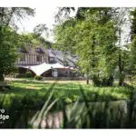 Cette carte postale de tero capture toute la beauté et l’authenticité du Tero Lodge Jalhay, un lieu d’exception niché au cœur de la nature. Entouré d’une forêt verdoyante et bordé par un étang paisible, ce magnifique bâtiment à colombages incarne le charme et l’élégance d’un refuge en pleine campagne. Une grande tente beige, installée en terrasse, offre un espace de détente et de convivialité pour profiter du cadre enchanteur. Loin du tumulte urbain, Tero Lodge invite à un séjour ressourçant, où gastronomie, bien-être et nature s’unissent pour une expérience inoubliable.