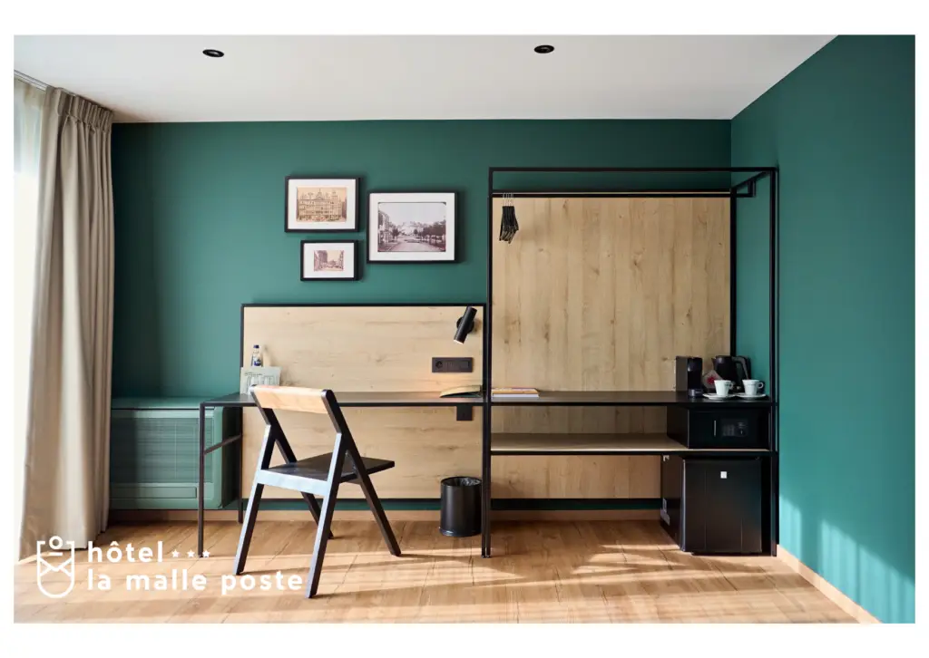 Cette carte postale de tero met en avant l’atmosphère élégante et apaisante d’une chambre de l’Hôtel La Malle Poste à Rochefort. La pièce est décorée avec soin, combinant des tons verts profonds, du bois clair et des éléments en métal noir pour un design à la fois moderne et chaleureux. Un bureau minimaliste en bois et métal est accompagné d’une chaise assortie, idéal pour travailler ou se détendre. Sur le mur, des cadres avec des photos d’époque rappellent l’histoire et le charme du lieu. Un coin café avec une machine et des tasses ajoute une touche de confort, tandis que la lumière naturelle traverse la pièce, accentuant son ambiance paisible. L’Hôtel La Malle Poste incarne un équilibre parfait entre authenticité et modernité, offrant un cadre idéal pour une escapade raffinée à Rochefort.