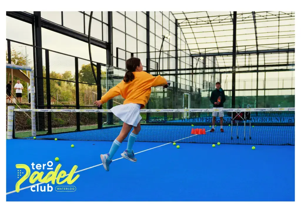 Cette carte postale met en avant l’énergie et la convivialité du Tero Padel Club Waterloo. Sur un terrain bleu éclatant, une jeune joueuse en jupe blanche et sweat orange effectue un smash avec détermination, incarnant l’esprit sportif et ludique du padel. En arrière-plan, un coach surveille l’entraînement, tandis que des balles jaunes parsèment le terrain, ajoutant du mouvement à la scène. L’environnement moderne et lumineux du club, entouré de baies vitrées, crée une atmosphère immersive et accueillante. Plus qu’un simple sport, le Tero Padel Club invite à un moment de partage et de dépassement de soi, accessible à tous, dans un cadre inspirant.